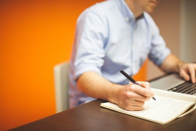 Man writing in notebook while looking at laptop 