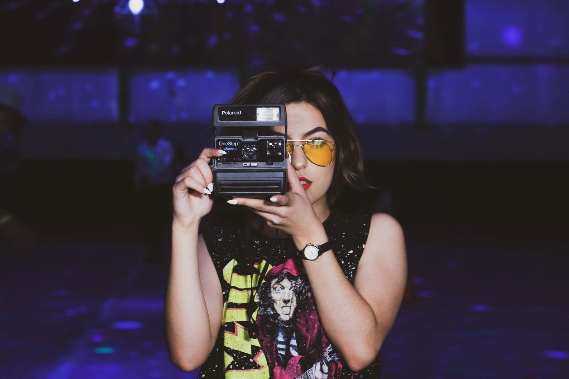 girl shooting black polaroid camera indoors against blue purple background